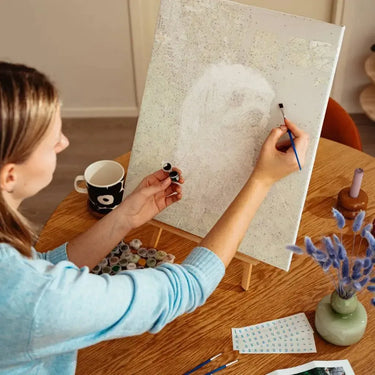 Nainen maalaa pöydän ääressä numeromaalausta, jossa näkyy koiran ääriviivat – paint by numbers -taulu työn alla.